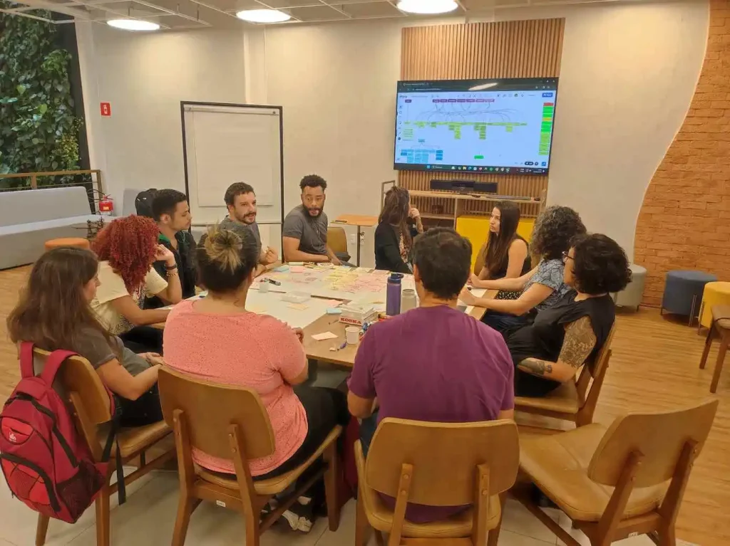 Imagem de um grupo de pessoas reunidas em workshop colaborativo com tela de televisão com gráfico ao fundo, ilustrando artigo sobre plataforma digital para síndico