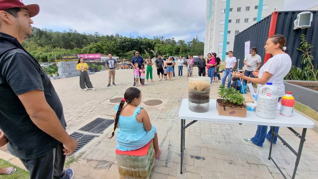 Grupo de moradores reunido em uma oficina de compostagem ao ar livre, com instrutora explicando o processo e demonstrando materiais, em atividade de ESG em condomínios