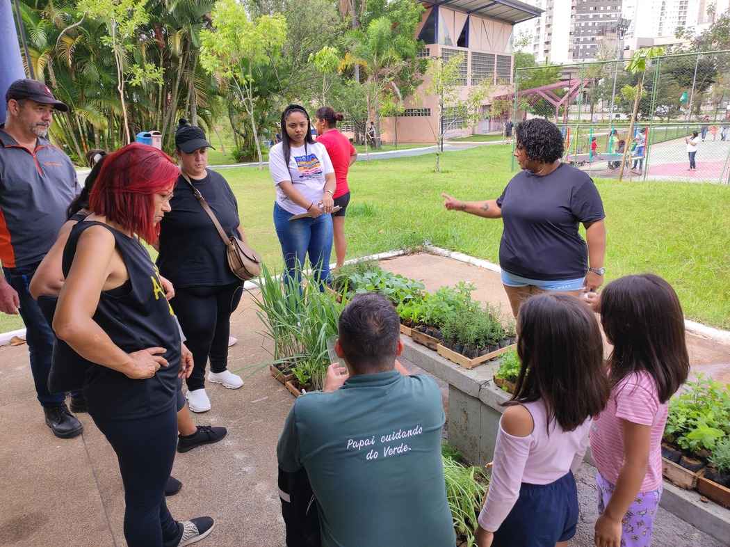 Grupo de moradores e crianças reunidos em uma oficina de plantio ao ar livre, aprendendo sobre hortas comunitárias como parte de ações de ESG em condomínios