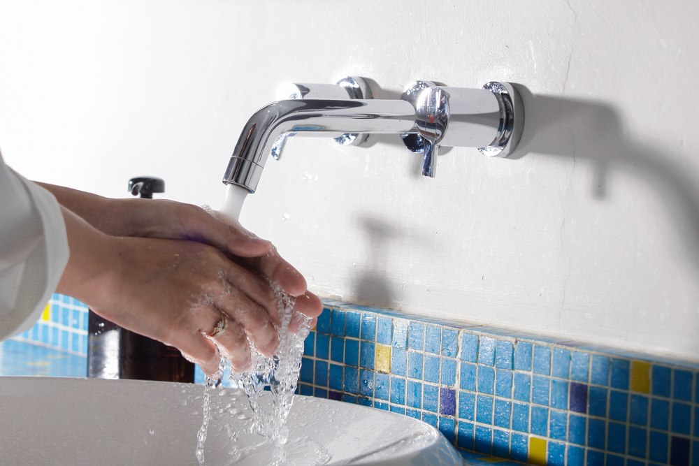 Mãos de uma pessoa sendo lavadas sob uma torneira aberta, com água corrente caindo sobre uma pia branca e detalhes de azulejos azuis ao fundo, ilustrando artigo sobre reajuste da tarifa de água da SABESP em 2026