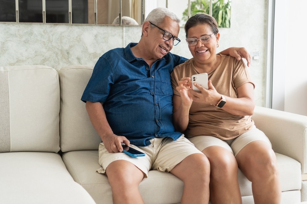 Casal sentado no sofá olhando para a tela de um celular, em momento descontraído dentro de casa, ilustrando artigo sobre regras de condomínio